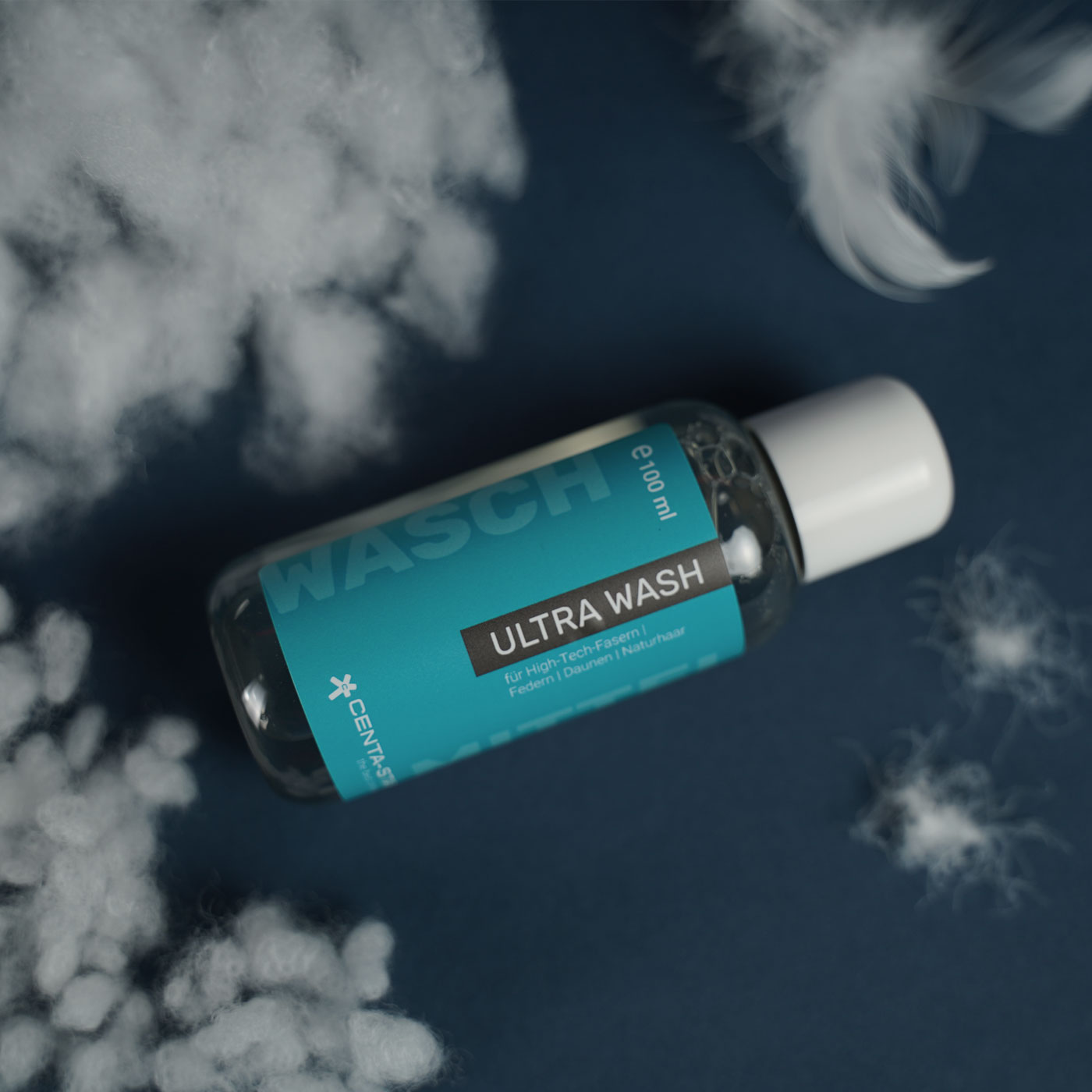 Centa-Star Ultra Wash Waschmittel – Das Waschmittel liegt vor blauem Hintergrund, seitlich sind Federn und Fasern platziert. Dies soll veranschaulichen, dass das Waschmittel für alle Materialien von Bettdecken und Kissen geeignet ist.